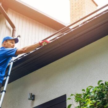 man on ladder cleaning house gutter from leaves and dirt