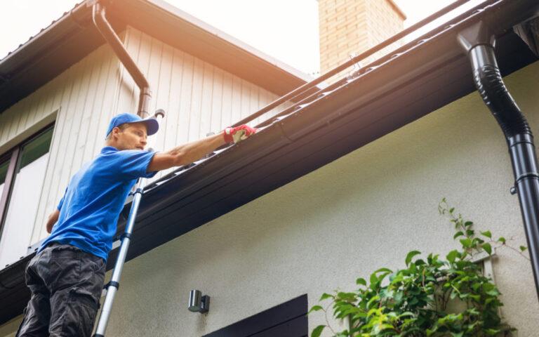 man on ladder cleaning house gutter from leaves and dirt